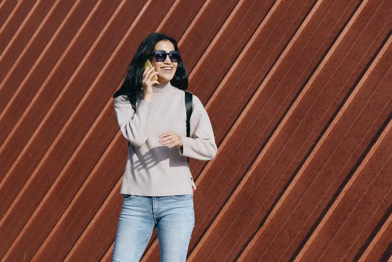 Woman wearing a sweater and jeans talks on a mobile phone while standing against a diagonal wood wall, blending casual fashion with modern urban ambience