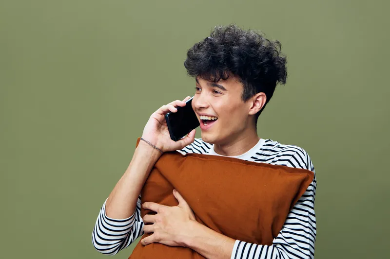 Smiling young man talking on smartphone while hugging a brown pillow, wearing striped shirt against green background, showing happiness and casual style.
