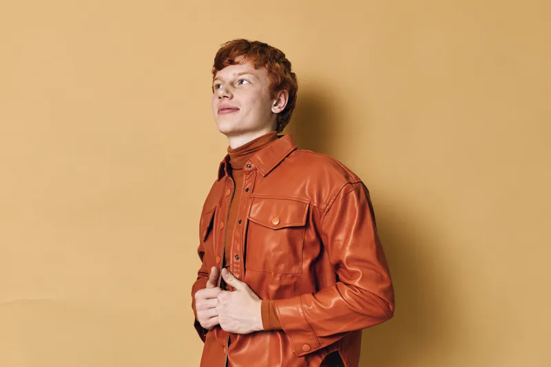Young man with red hair wearing a brown leather jacket standing against beige background looking away with confident expression and casual style in studio portrait.