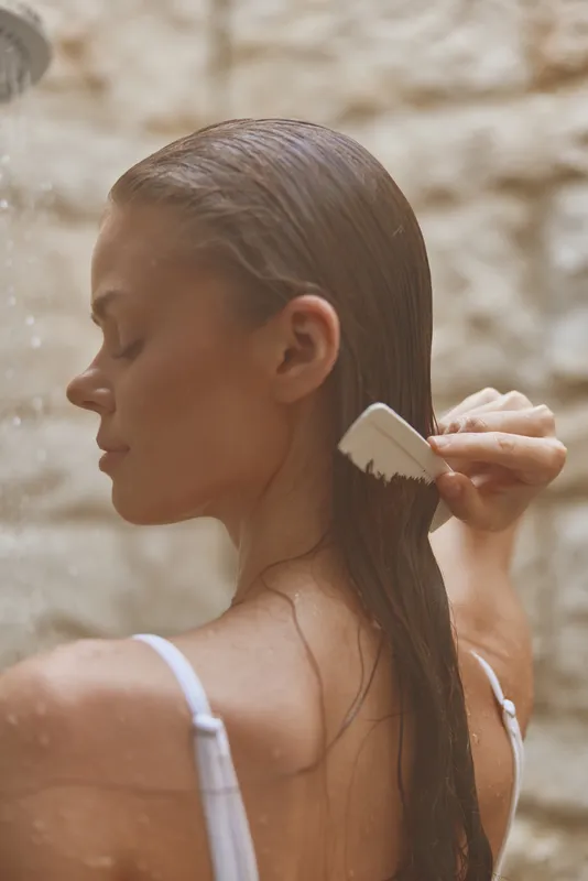 Woman brushing wet hair under soft natural light, showing calm and relaxation with blurred warm background tones, captured in intimate side profile.