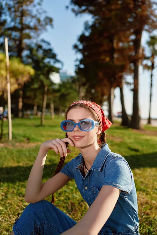 young woman wearing blue sunglasses and red headscarf sitting on grass in sunny park surrounded by tall trees and greenery