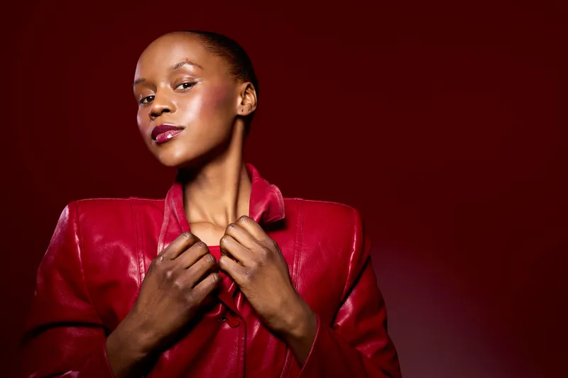 Stylish African woman with dark skin wearing a red leather jacket poses confidently against a red background. Fashion portrait highlighting elegance and bold color contrast.