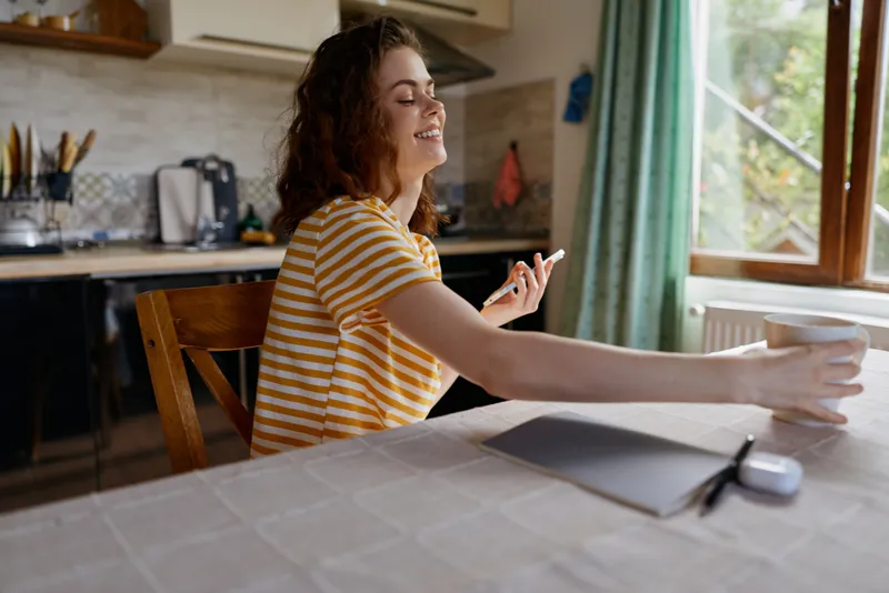 A smiling woman in a striped shirt sits at a kitchen table, enjoying a casual moment at home. Bright daylight streams in as she reaches for a mug, capturing everyday comfort.