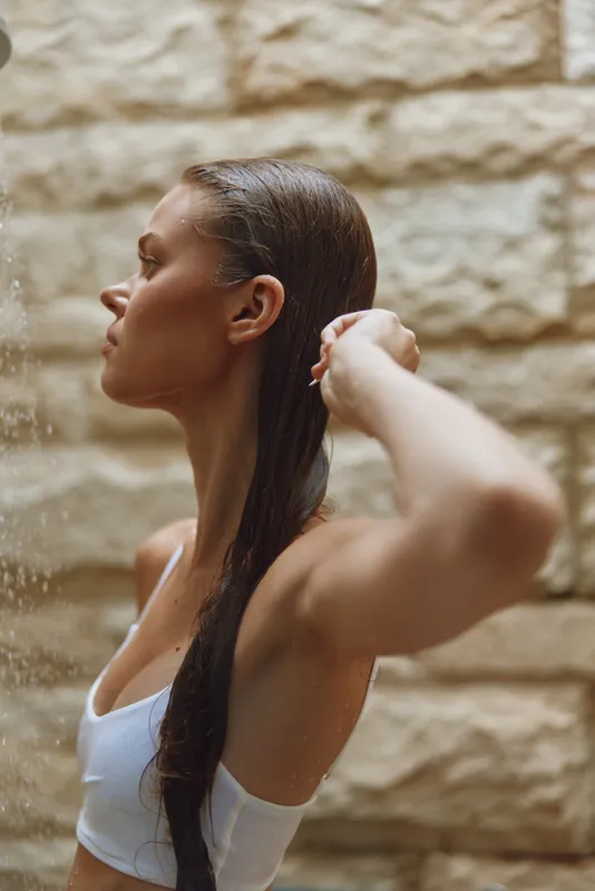 Young woman washing her long wet hair under a warm shower with natural stone wall background in a peaceful indoor bathroom setting.