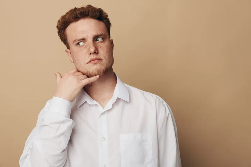 Young man with red hair and freckles in white shirt makes call me gesture with hand near face, looking sideways on beige background. Concept of communication, gesture, and casual style.