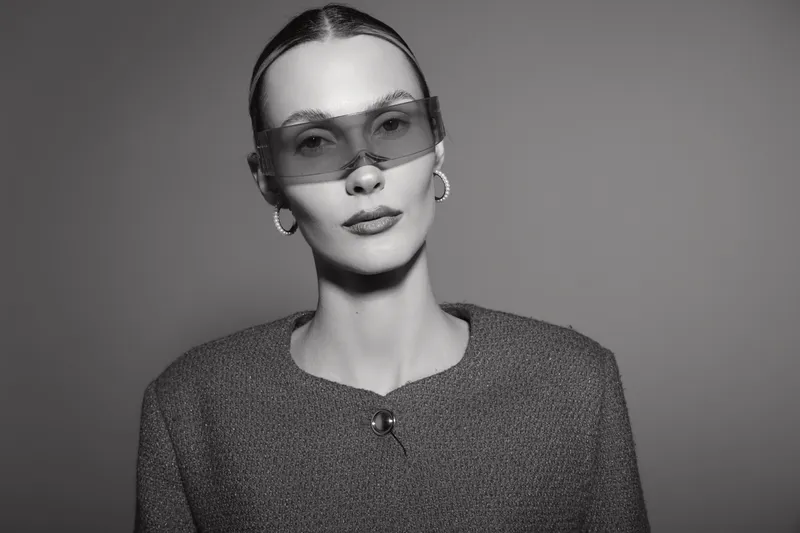Elegant white woman with fair skin and slicked hair wearing oversized sunglasses and textured jacket in studio.
