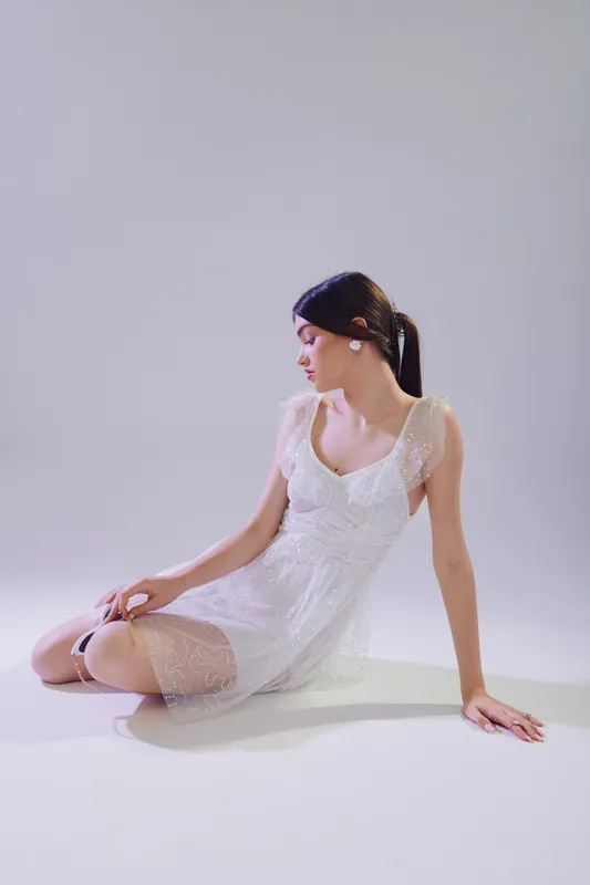 Young woman in a white delicate dress sits on the floor looking sideways. Soft lighting emphasizes her beauty and fashion style. Concept of elegance, femininity, and modern trends.
