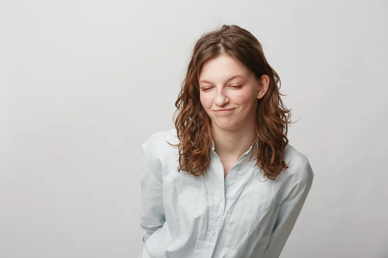 Young woman with curly brown hair in white shirt looks down with slight smile and closed eyes. Studio portrait on plain light background. Concept of emotion, shyness, and introspection.