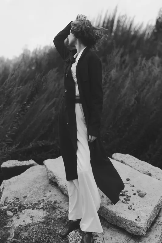 Fashion in motion on a windy coastline, long coat over a flowing dress, monochrome scene with rugged rocks and tall grasses, expressive movement and dramatic atmosphere