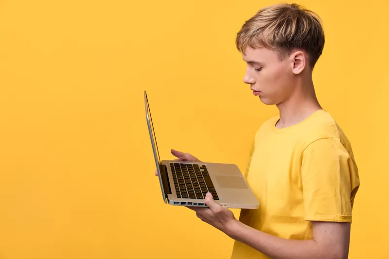 Young man in a yellow t shirt holds a laptop against a bright yellow background, showcasing digital work, technology exploration and creative learning in a modern setting