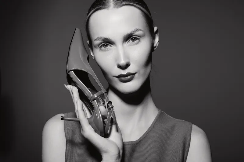 Elegant Caucasian woman with fair skin and slicked back hair holds high heel shoe near face. Studio portrait in black and white with minimalistic background and fashion concept.