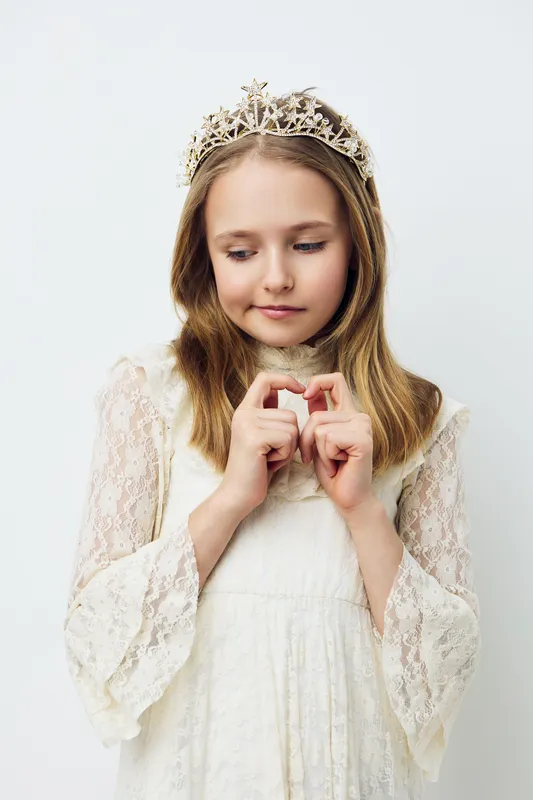 A young girl in a white lace dress with a crown, expressing innocence and wonder against a soft white background, perfect for themes of childhood and elegance