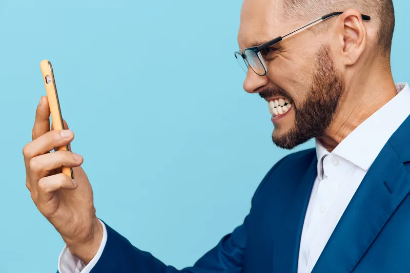 A professional man in a suit stands against a solid blue background, looking at a smartphone with a confident, focused expression, conveying ambition and expertise in a business setting.