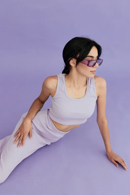 Young woman in lavender outfit and purple sunglasses sitting on pastel purple background, showcasing stylish casual fashion with relaxed pose and modern look.