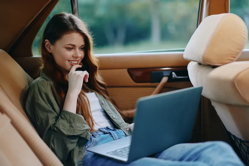 Young woman in casual attire, smiling while using a laptop in a car, showcasing a relaxed and productive mood in a natural setting.