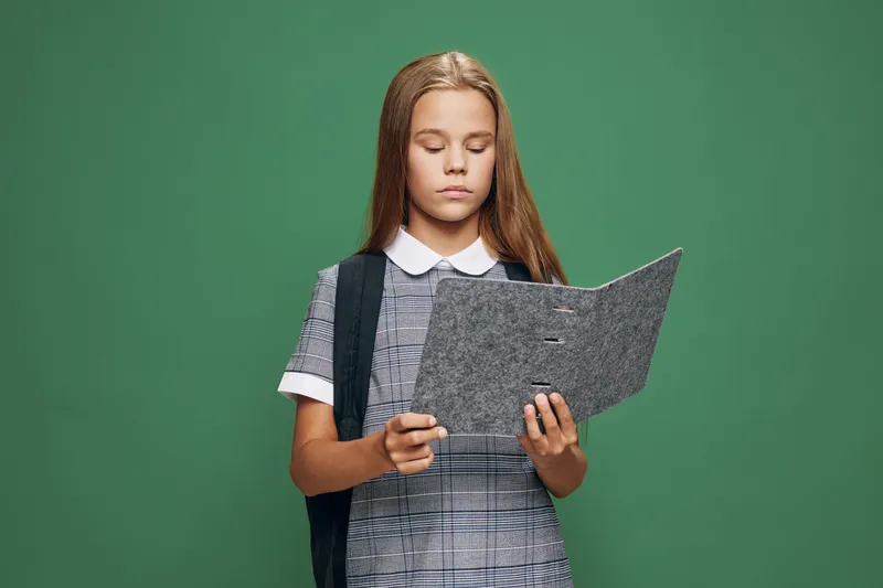 Girl student with backpack reading folder on green background for education, school, learning and study concept with focused expression and uniform outfit.