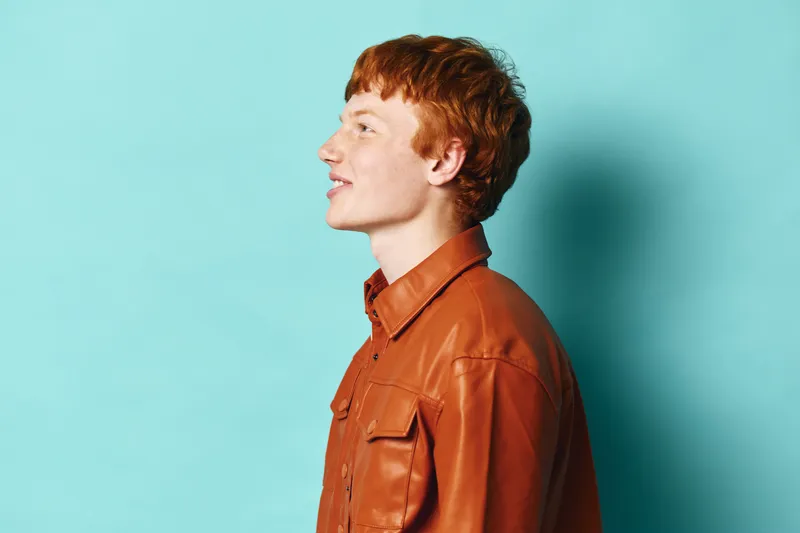 Young man with red hair wearing brown leather jacket posing in profile against blue background for fashion portrait and style concept.