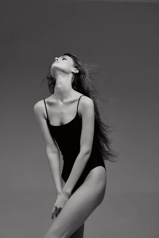 Black and white portrait of a slender woman in a black swimsuit posing elegantly against a monochrome background.