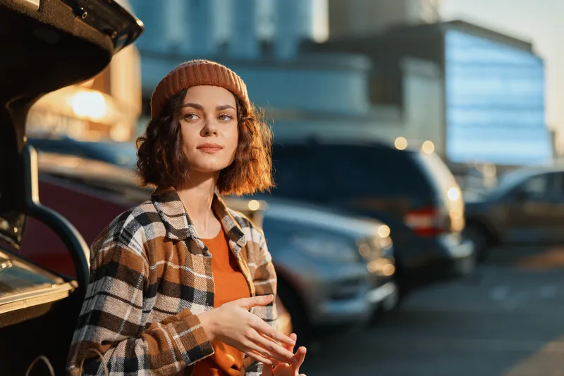 Woman portrait holding coffee in urban parking at golden hour, candid authenticity and mindful living vibe. Emotional storytelling, cozy beanie, plaid coat, modern city lifestyle.