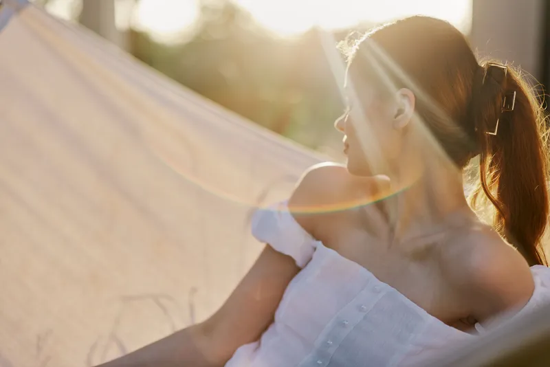 Young woman relaxing in a hammock during sunset, wearing a light summer dress, exuding tranquility and joy, surrounded by nature, with warm colors and soft light
