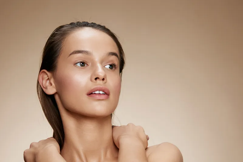 Beautiful young woman with natural makeup and healthy skin, expressing confidence against a soft beige background showcasing beauty and wellness