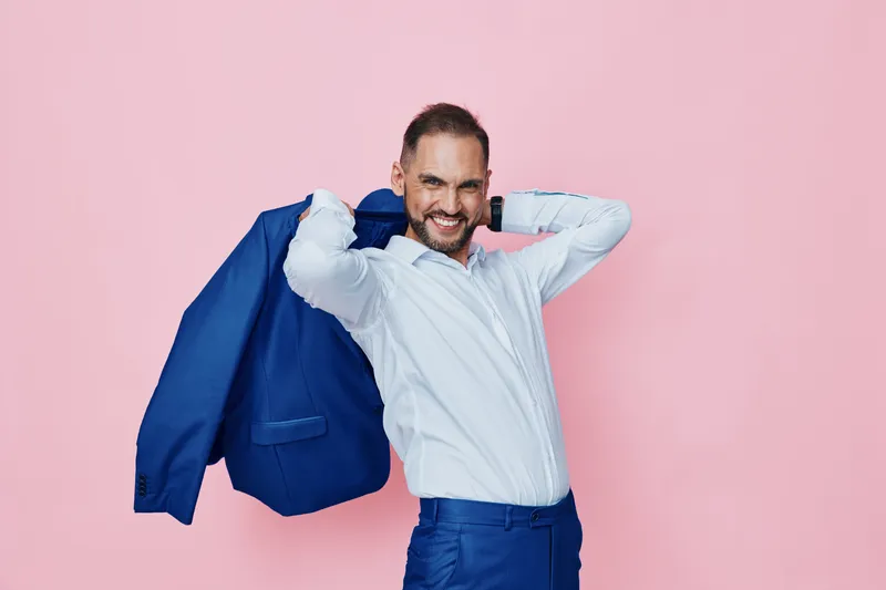 A professional man exudes confidence on a pink studio backdrop, wearing a crisp white shirt and blue trousers, draping a blue blazer over his shoulder with a bold smile and poised stance.