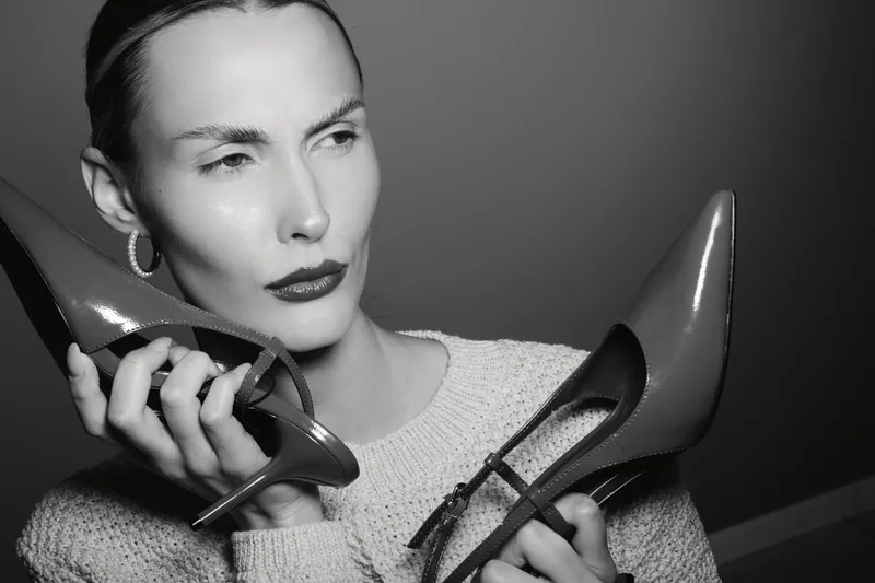 White woman with pale skin holds two high heel shoes near face, looking confused. Concept for fashion choice, style, and decision making.