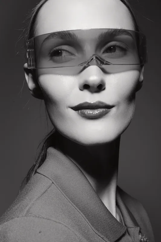 Portrait of a Caucasian woman with fair skin wearing transparent futuristic glasses and a collared jacket in black and white studio lighting.
