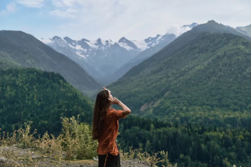 A woman stands on a hillside gazing at snow capped mountains in a lush valley, capturing a tranquil outdoor landscape and wide open nature