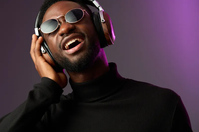 Young Black man enjoying music with headphones, wearing sunglasses, vibrant background, emotional expression of happiness and freedom, modern lifestyle