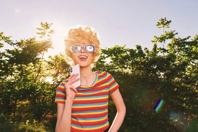 Bright woman with retro style wears striped shirt and sunglasses, standing outdoors in warm sunlight, capturing a playful lifestyle image with vibrant, cheerful energy.