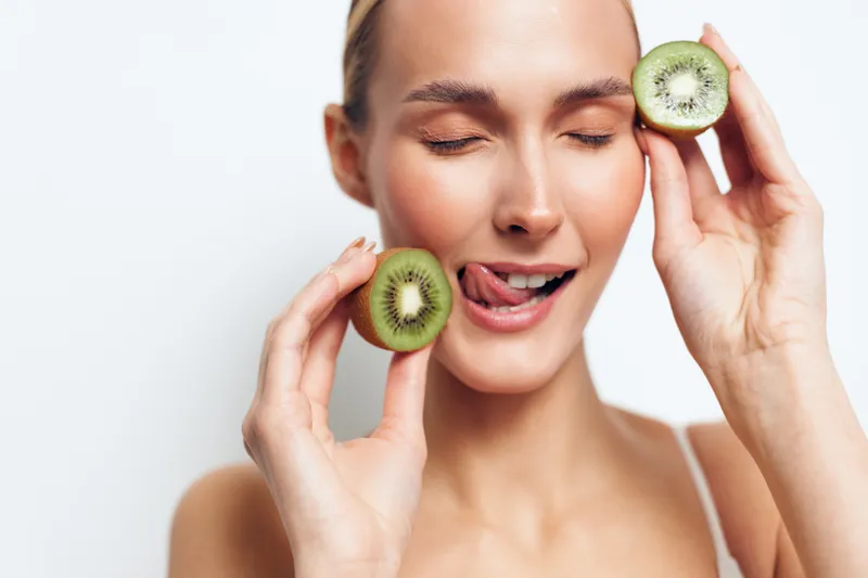 Woman holding kiwi slices to eyes, skincare spa wellness fruit mask concept, smiling face closed eyes, beauty treatment on white background, soft gentle studio light