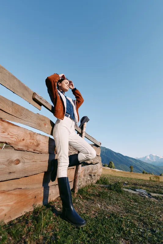 Woman by wooden fence in mountains wearing boots, sweater and sunglasses, posing in countryside meadow under clear sky. Fashion portrait, outdoor lifestyle, travel and nature.