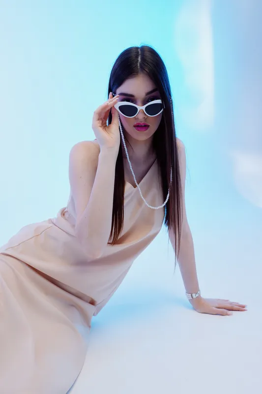 Young woman in a beige dress poses wearing white sunglasses with a pearl chain. Her long dark hair is straight. Studio shot with soft blue lighting. Concept of beauty fashion elegance.