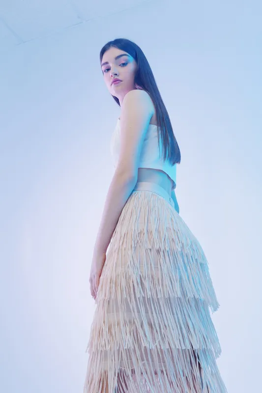Young woman with long hair wearing a fringed skirt and sleeveless top poses against a soft blue background. Concept of woman beauty fashion and modern style.