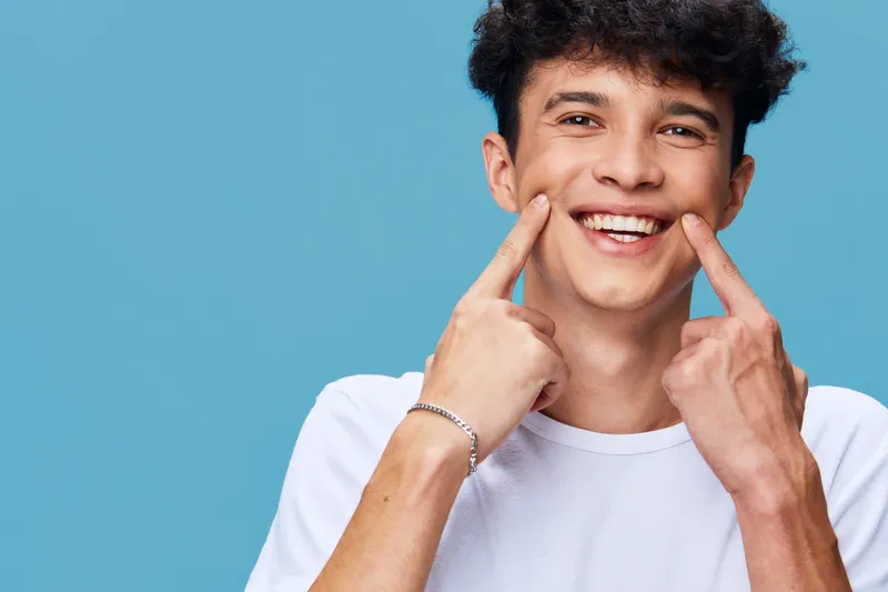 Smiling man pointing at cheeks with fingers, happy young adult male wearing white t-shirt against blue background, positive emotion and casual style portrait.