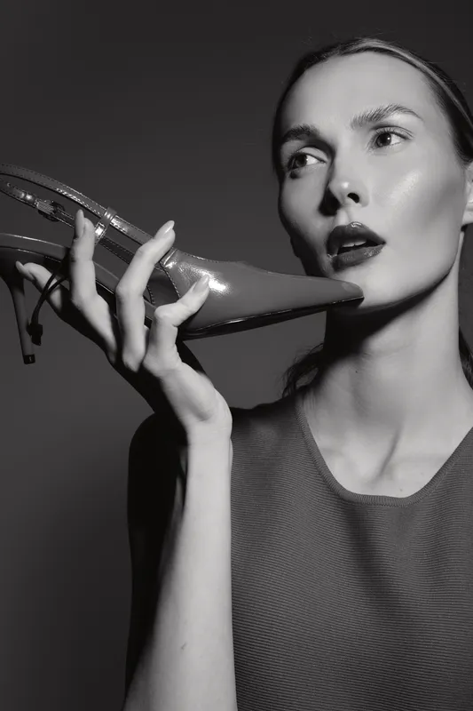 Caucasian woman with light skin holds a high heel shoe near her face, looking thoughtful in a black and white studio portrait. Concept of fashion and elegance.