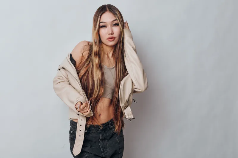 Young woman with long hair wearing casual jacket and crop top posing against gray wall. Fashion, beauty, style and confidence in studio portrait of female model with natural makeup and trendy outfit.