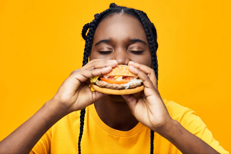 Young Black woman enjoying a delicious burger against a vibrant yellow background, radiating happiness and excitement in a casual yellow t shirt