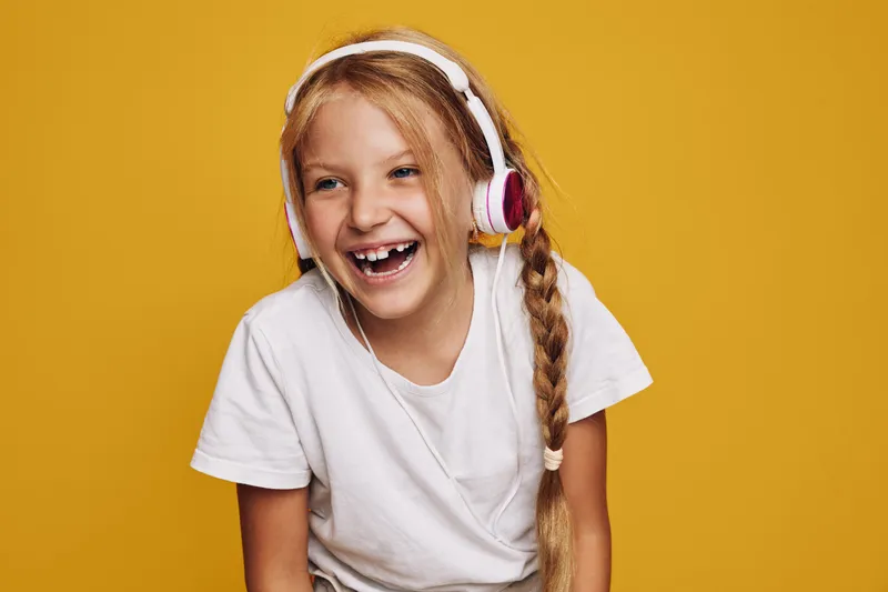 Happy girl with long braided hair wearing large headphones laughs joyfully against a yellow background. Casual white t-shirt and positive energy. Concept for childhood and music.