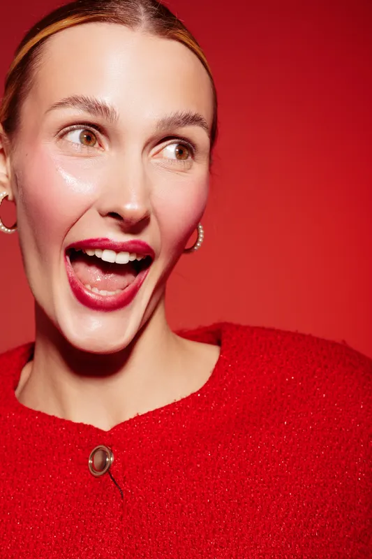 Excited young woman with bright red lipstick and red sweater smiles widely against a red background. Concept of happiness, fashion, beauty, and vibrant emotions.