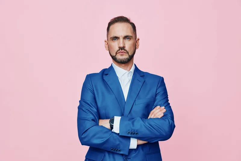 A confident business professional stands with arms crossed against a soft pink backdrop, portraying focus, determination, and poised leadership in a modern work setting.
