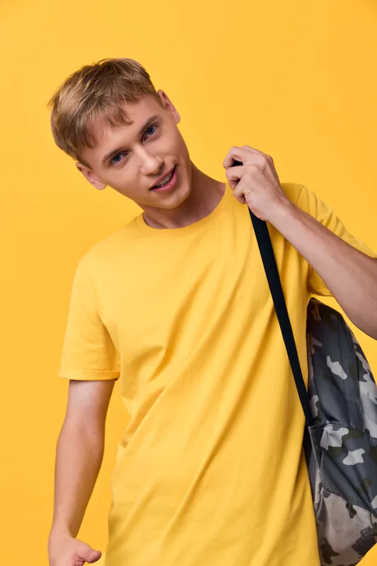 Young man in a yellow t shirt with a camo backpack strap over his shoulder against a bright yellow background, expressing casual lifestyle fashion and mobility