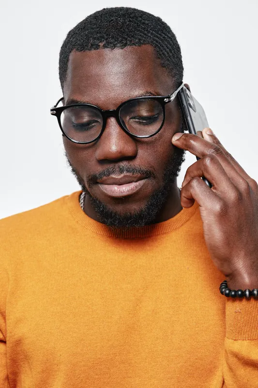 African man with dark skin and glasses talking on smartphone wearing orange sweater. Closeup portrait of thoughtful black male using mobile phone for communication and technology.