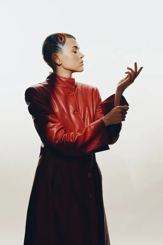 Woman with colorful hair wearing a red leather coat poses gracefully against a minimalist white background showcasing creative style and bold fashion.