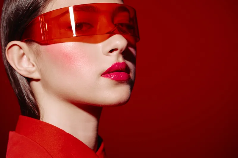 Close-up portrait of a fashion model with bright makeup and glossy lips wearing futuristic red translucent glasses on red background. Beauty and creative style.