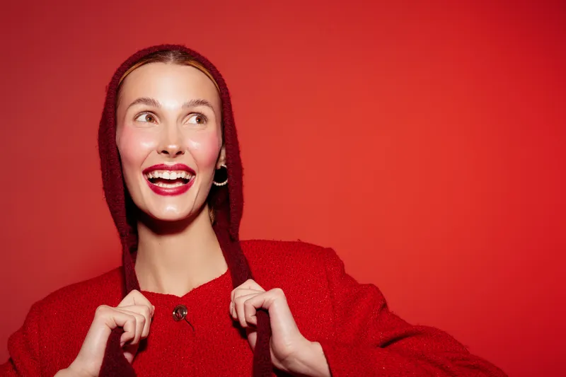 Smiling young woman in red sweater with hood looks away on red background. Bright makeup and joyful expression create lively and festive mood. Concept of happiness and fashion style.