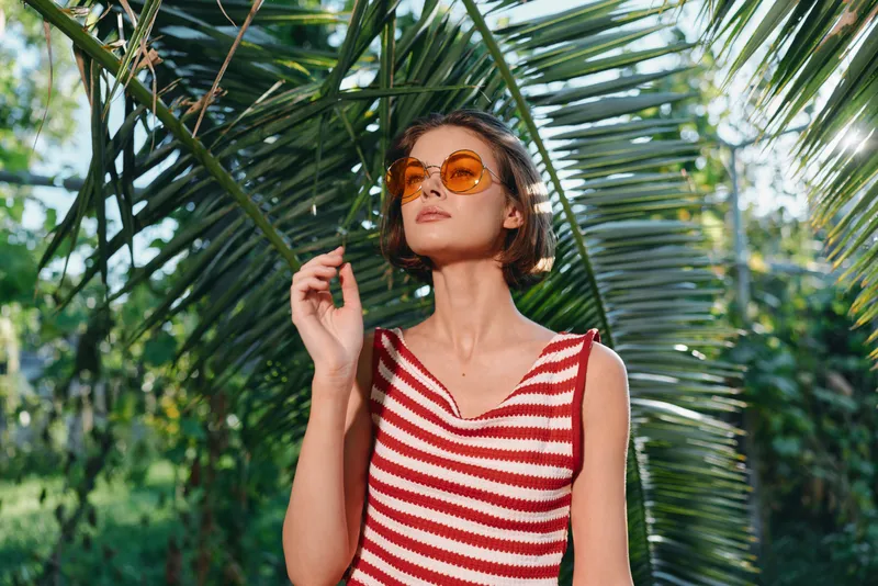 A stylish woman stands amid lush tropical foliage, wearing a red and white striped sleeveless top and round orange sunglasses, gazing upward with a relaxed, sunlit vibe.