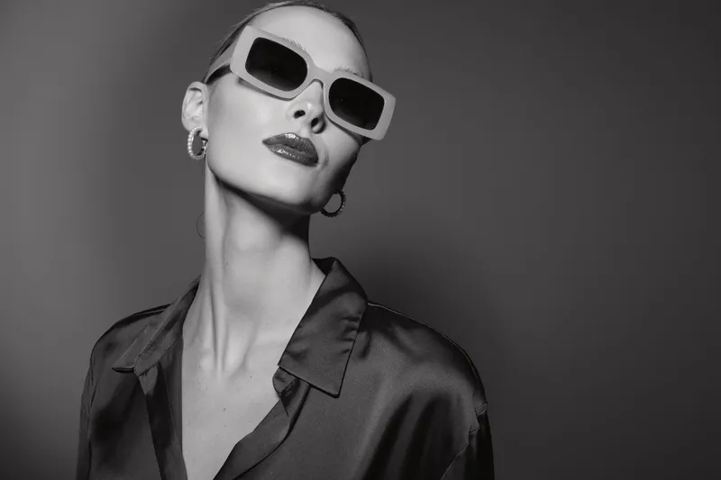 Stylish white woman with pale skin wearing sunglasses and silk shirt poses confidently against dark background in black and white.