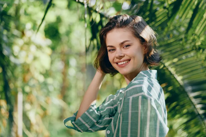 Smiling woman in striped shirt stands in lush outdoor setting with natural light and a carefree portrait vibe.